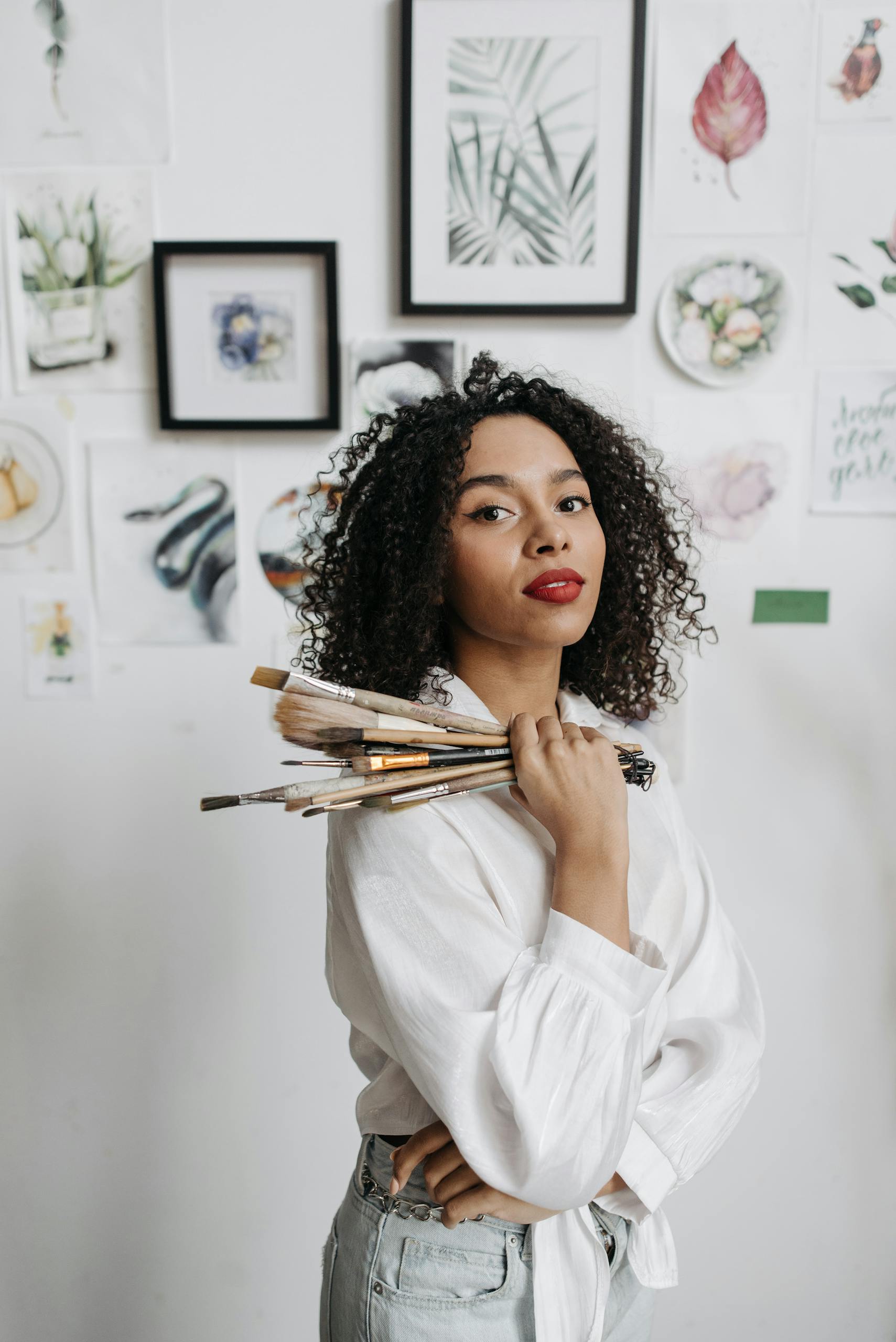 Elegant woman with curly hair holds paintbrushes in art studio against a backdrop of framed paintings.
